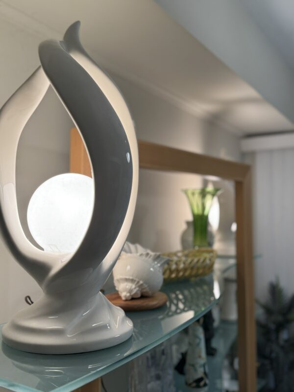 A modern, sculptural white lamp with a glowing orb sits on a glass shelf. Behind it are decorative items, including a green vase with flowers, a seashell, and various ceramics, all illuminated by soft indoor lighting.