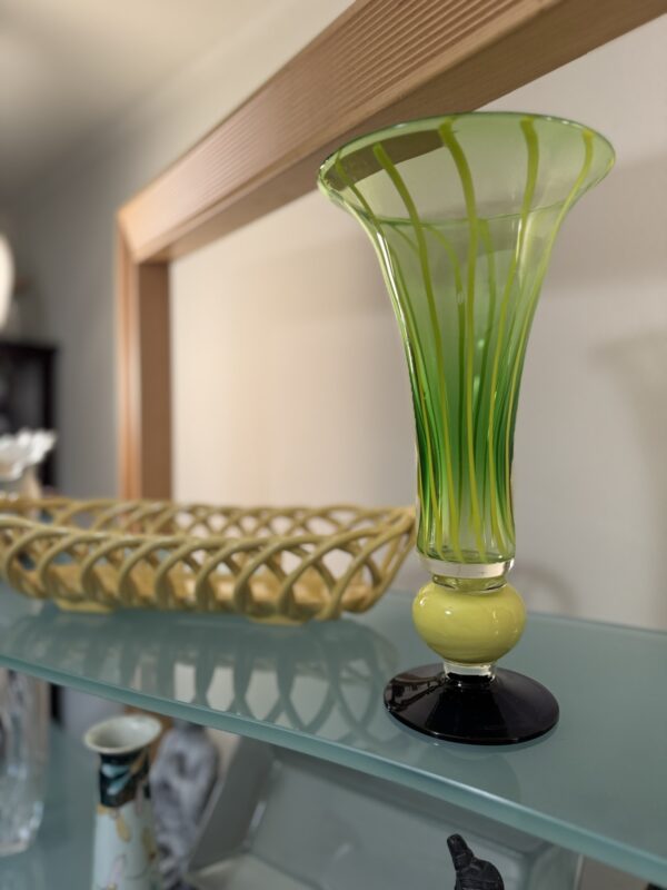 A green and yellow glass vase with a wide flared top and a black base stands on a glass shelf. Behind it is a tan woven basket. The background shows a softly lit room and parts of other glassware out of focus.