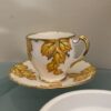 A small white teacup and saucer decorated with yellow and gold leaf patterns sit on a glass surface. The cup has a gold-rimmed handle, and part of a white ashtray is visible in the foreground. A plain beige wall is in the background.