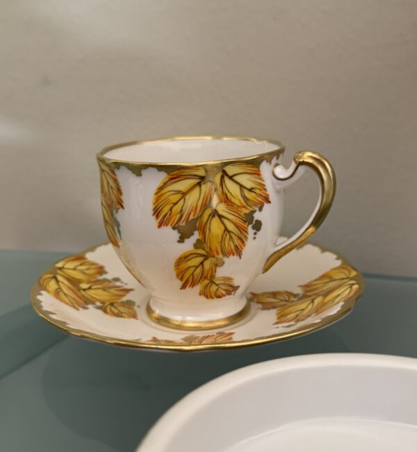 A small white teacup and saucer decorated with yellow and gold leaf patterns sit on a glass surface. The cup has a gold-rimmed handle, and part of a white ashtray is visible in the foreground. A plain beige wall is in the background.