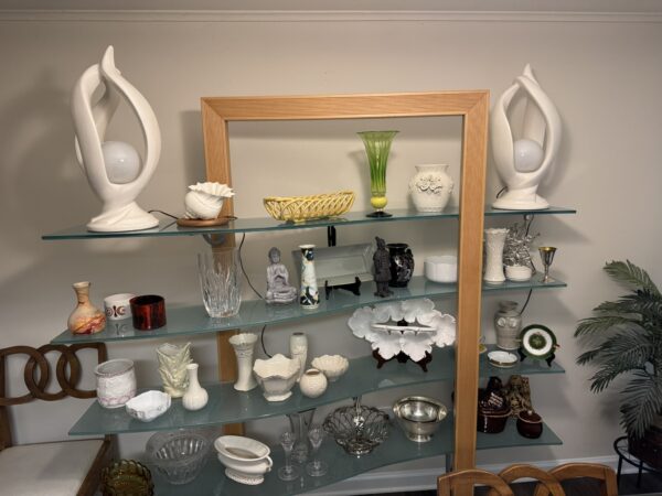 A glass display shelf holds assorted decorative items: white abstract lamps on top, colorful vases, glass bowls, ceramic figurines, ornate dishes, and small collectibles. A potted plant is on the right and wooden chair backs are visible on the left and bottom.