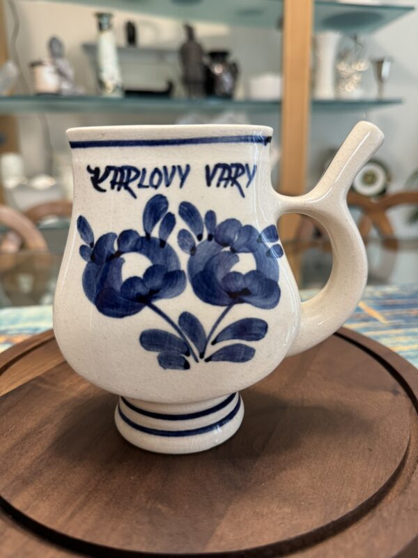 A ceramic mug with a pipe-like spout, decorated with blue floral patterns and KARLOVY VARY written in blue near the rim, sits on a wooden tray. Shelves with glassware and ceramics are visible in the blurred background.