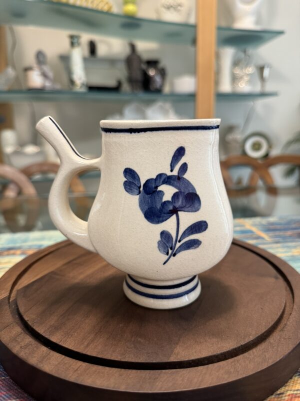 A ceramic mug with a handle designed for easy gripping sits on a round wooden tray. The mug features a blue floral pattern on white glaze, with dark blue trim at the rim and base. In the background, shelves display decor and glassware.
