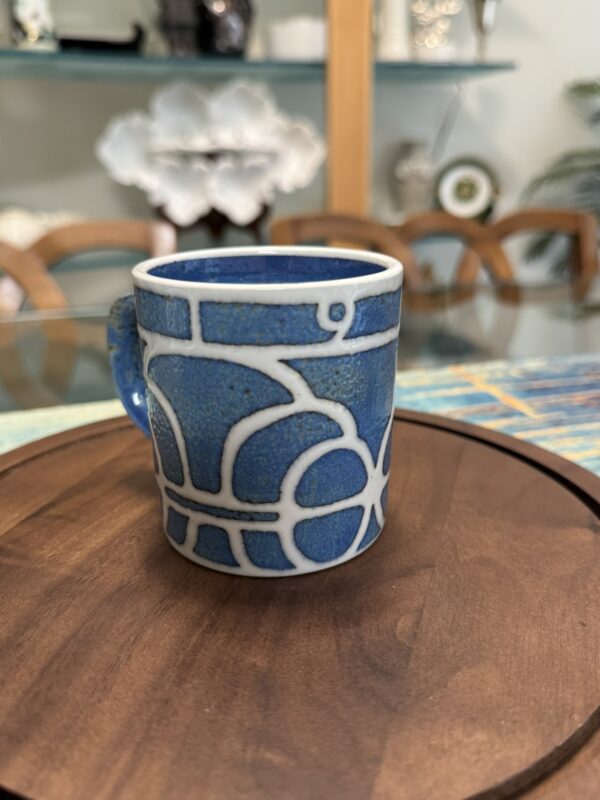 A small blue ceramic mug with a white abstract geometric pattern sits on a round wooden tray. In the background, there is a glass table, wooden chairs, a striped placemat, and decorative shelves with white ceramic items.