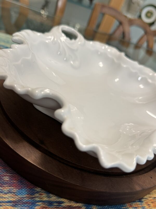 A close-up of a decorative white porcelain dish with ornate, ruffled edges and a small handle, resting on a round wooden tray atop a colorful woven tablecloth. In the blurred background, there are chairs and shelves with dishes.
