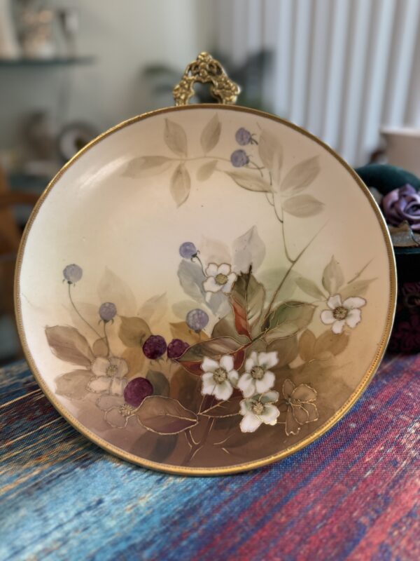 A decorative ceramic plate with a gold rim features a floral and berry design. White flowers with yellow centers and green leaves are painted alongside purple and blue berries. The plate stands upright on a table with a colorful woven cloth.