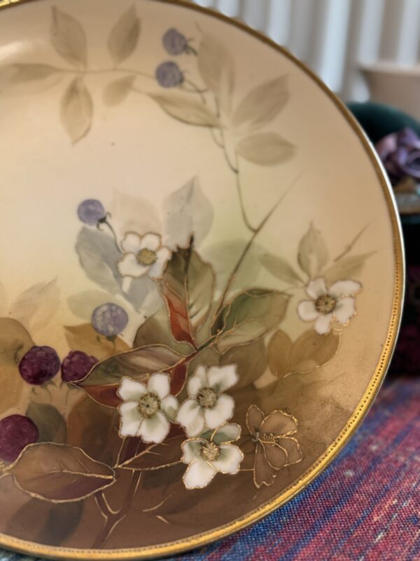 A decorative plate with a gold rim features a painted design of white flowers, green leaves, and purple berries. The background is a soft beige, and the plate rests on a colorful, textured fabric. The scene has a delicate, vintage style.