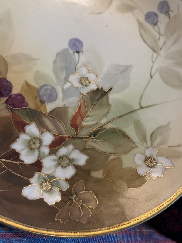 A close-up of a decorative plate featuring white flowers with gold outlines, green and brown leaves, and small purple berries on a soft, pale background. The plate’s edge is gold, and some bright blue and purple fabric is visible underneath.
