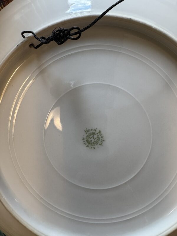 The underside of a white ceramic plate with a green mark reading Hand Painted Nippon around a wreath logo at the center; a black string is loosely knotted and partially resting on the plate’s rim.