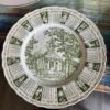 A decorative plate displays a green illustration of a church with trees and the words God Bless Our Home Throughout 1965. Around the edge, each month of 1965’s calendar is shown in boxes with zodiac symbols, all in green on a white background.