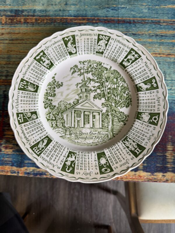 A decorative plate with a green illustration of a church and trees in the center, reading God Bless Our House Throughout 1965. The rim features the 1965 calendar months and zodiac symbols in green, set against a patterned, multicolored tablecloth.