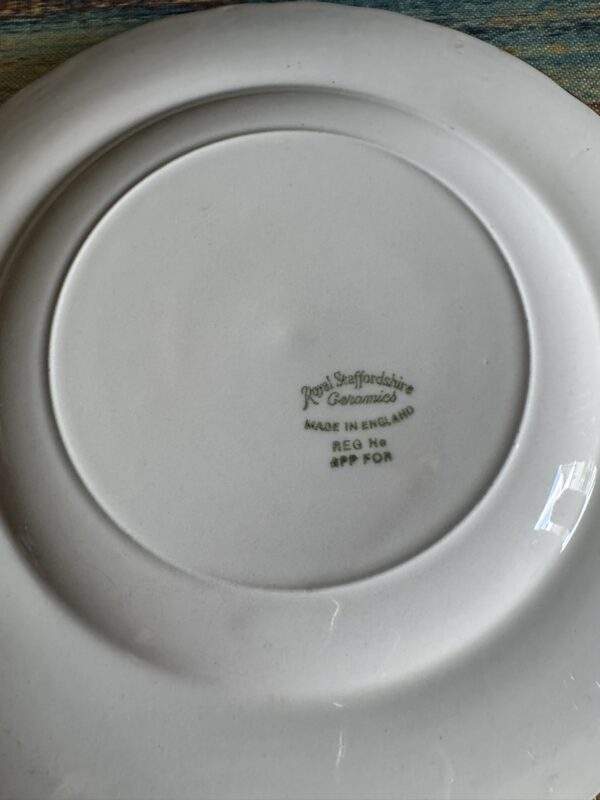The underside of a white ceramic plate shows a green stamp that reads: Royal Staffordshire Ceramics Made in England Reg No 879 For. The plate is resting on a textured blue and yellow surface. The lighting casts a faint reflection on the plate.