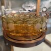 A vintage amber glass bowl with a scalloped rim sits on a round wooden base. Decorative etched coins or medallions encircle the bowl. In the background, glass shelves display various white and clear decorative objects and ceramics.