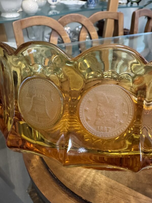 An amber-colored glass bowl with a wavy rim features embossed designs: the Liberty Bell on one side and a U.S. coin dated 1877 on the other. The bowl sits on a wooden table with chairs and ceramic items blurred in the background.