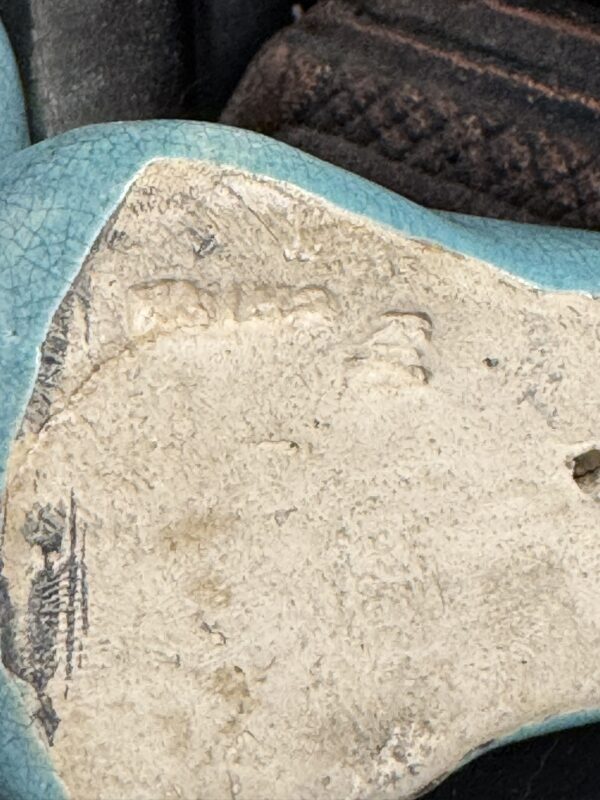 Close-up of the base of a turquoise-glazed ceramic object showing worn and partially illegible maker’s mark or inscription on the rough, unglazed underside.