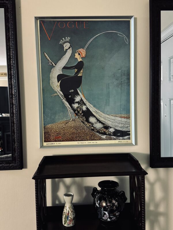 Framed vintage Vogue magazine cover featuring an elegant woman in a black dress holding a white peacock, hanging above a dark wooden shelf with a black vase and a small decorative bottle.
