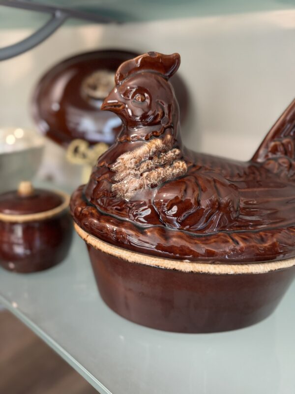 A glossy, brown ceramic dish shaped like a hen sits on a glass shelf. The lid forms the hens body. In the background, smaller matching brown ceramic containers and a large oval platter are also visible. The scene is softly lit and slightly blurred.