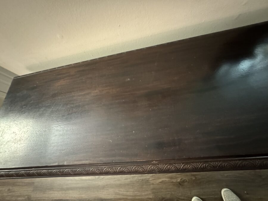 A dark wooden tabletop with a glossy finish showing some scuffs and light scratches. The table’s edge has a carved pattern, and part of a wooden floor and white shoes are visible at the bottom right corner. Neutral wall in the background.