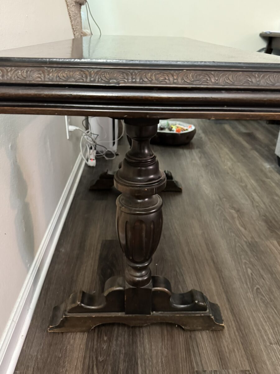 A dark wooden table with ornate carvings on the edge, featuring a single central pedestal leg with decorative details. The table stands on a dark hardwood floor near a light-colored wall with visible baseboards and power cords.