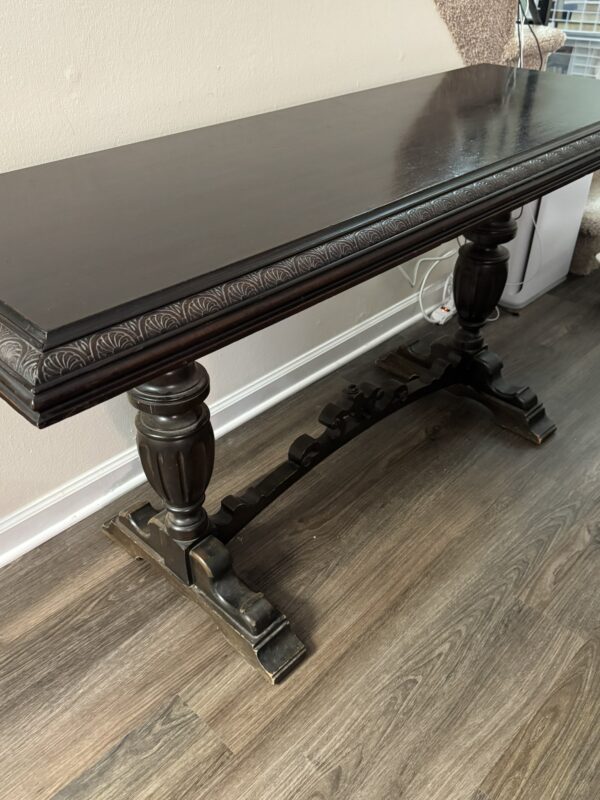 A dark wooden console table with ornate carved edges and two thick, turned legs connected by a decorative stretcher. The table sits on a textured wood floor near a wall and carpeted stairs. Visible power cords are tucked behind the table.