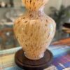 A large, yellow and brown speckled glass vase with a wide, flared rim sits on a round dark wooden base. The vase is displayed on a colorful woven table runner, with blurred shelves and a plant visible in the background.
