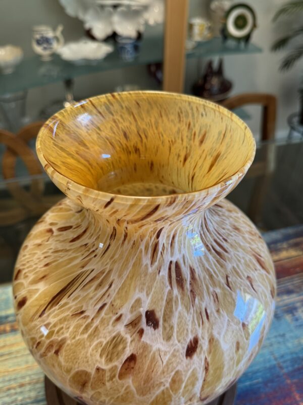 A close-up of a wide-mouthed, hand-blown glass vase with a mottled pattern of golden yellow, amber, and brown. The vase sits on a table with a colorful cloth. In the background, there are wooden chairs and glass shelves displaying decorative ceramics.