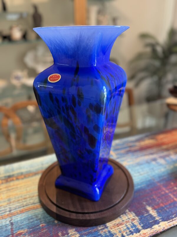 A tall, square-shaped cobalt blue glass vase with dark speckled patterns sits on a round wooden base atop a colorful, woven tablecloth. The vase has a flared rim, and a sticker near the top reads Murano. Blurred shelves and plants are in the background.