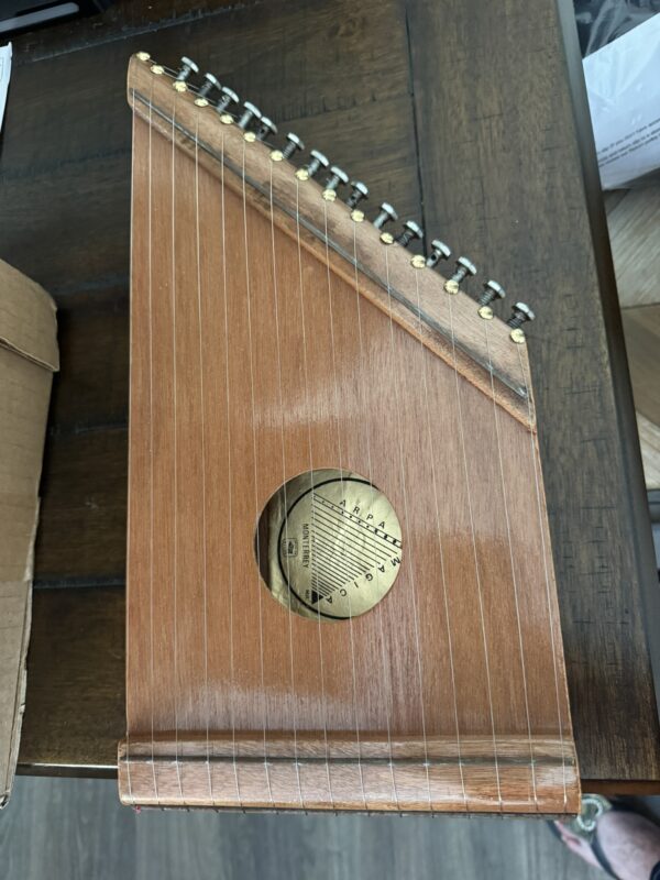 A wooden autoharp with a trapezoidal shape sits on a dark wooden table. The instrument has metal tuning pegs along one edge, twelve strings, and a round sound hole with a gold label featuring the note names for tuning. Part of a cardboard box is visible on the left.