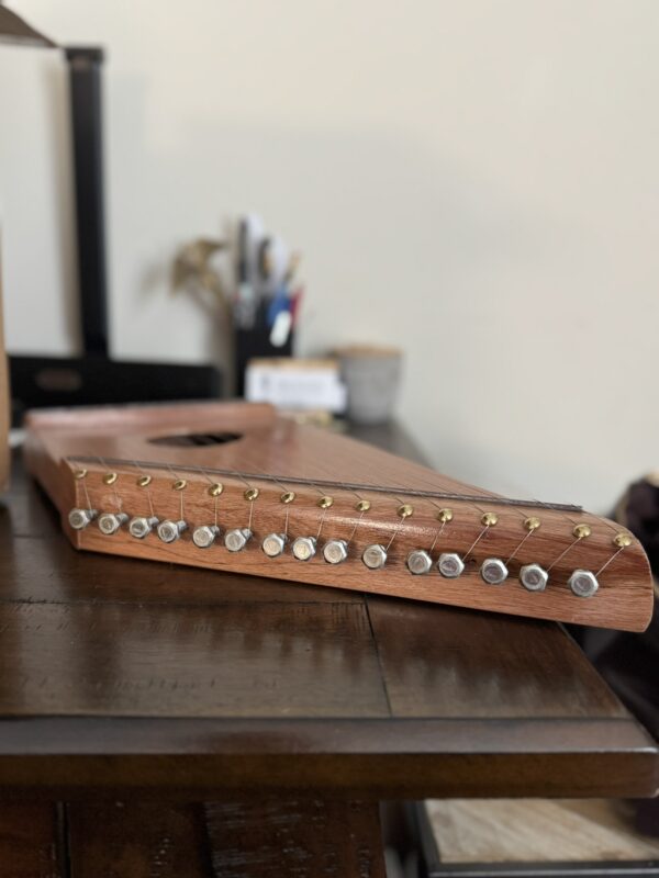 A wooden stringed instrument, likely a zither or autoharp, rests on a dark wooden table. The instrument has tuning pegs with round silver heads and golden tuning pins. The background is softly blurred, showing pens and other desk items.