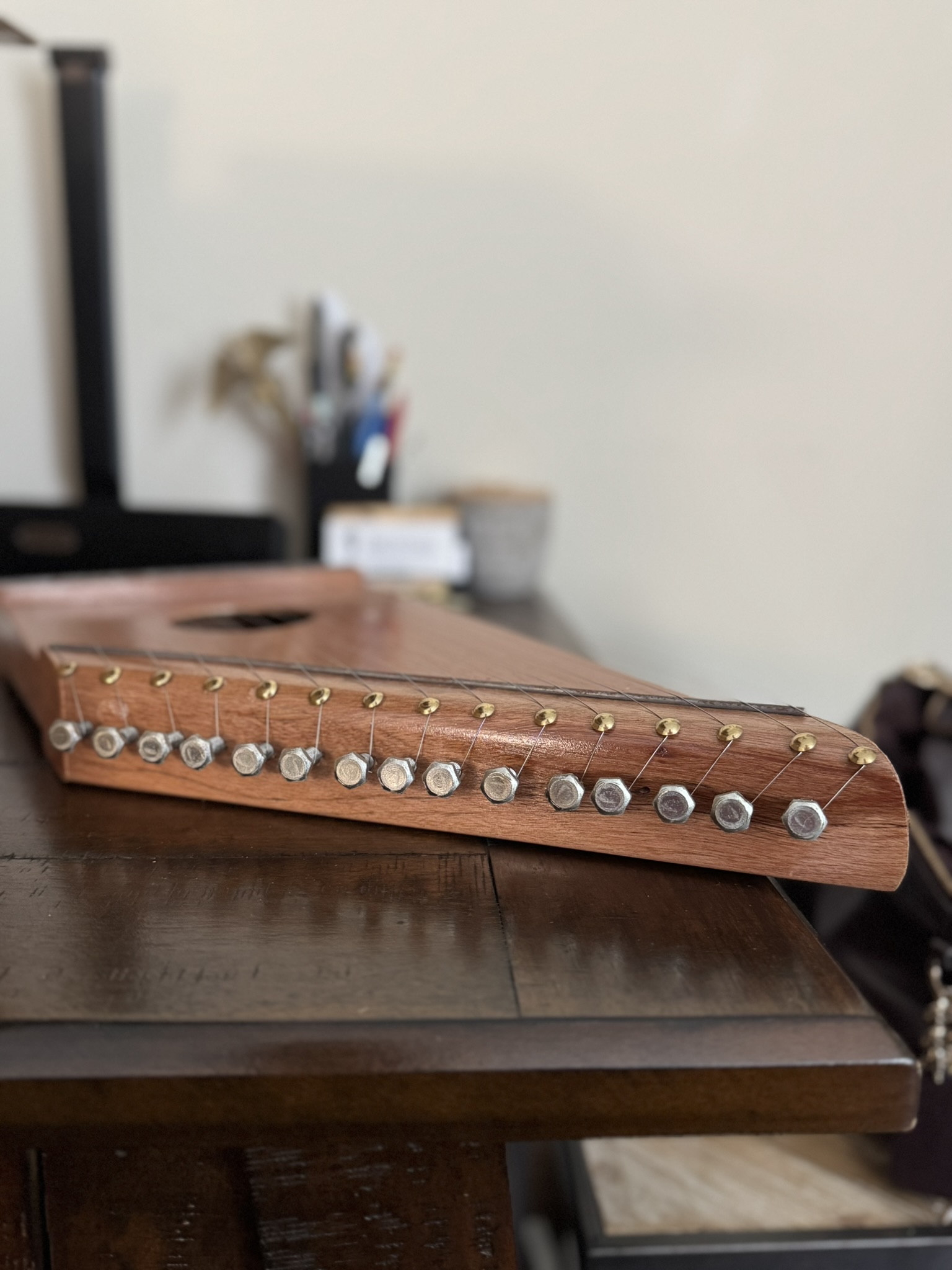 A wooden lap harp with metal tuning pins and strings sits on a dark wooden table. Office supplies and a blurred desk are visible in the background. The image is focused on the harp’s strings and tuning pins, highlighting its craftsmanship.