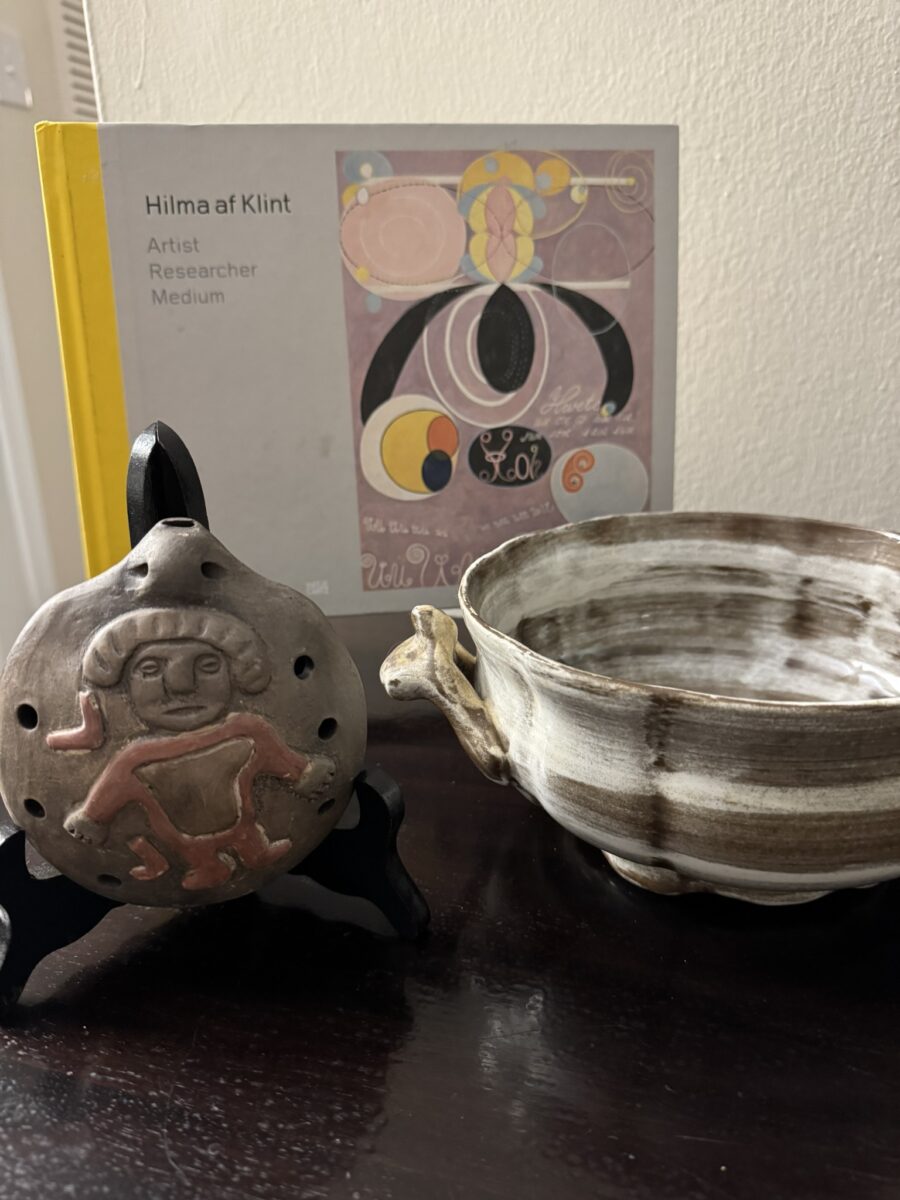 A round, brown ceramic piece with a face and red accents stands on a black holder beside a marbled gray and white bowl with a small handle. Behind them is a book titled Hilma af Klint: Artist Researcher Medium, partly open to a colorful artwork.