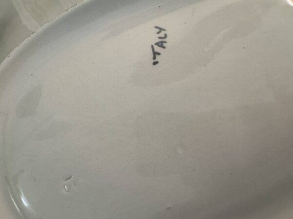 Close-up of the bottom of a white ceramic plate with the word “ITALY” handwritten in black ink near the center. The surface shows some minor water spots and faint scratches, and there are no visible maker’s marks besides the writing.