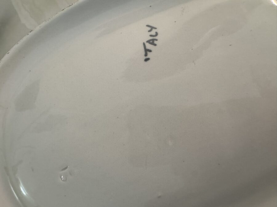 Close-up of the bottom of a white ceramic plate with the word “ITALY” handwritten in black ink near the center. The surface shows some minor water spots and faint scratches, and there are no visible maker’s marks besides the writing.