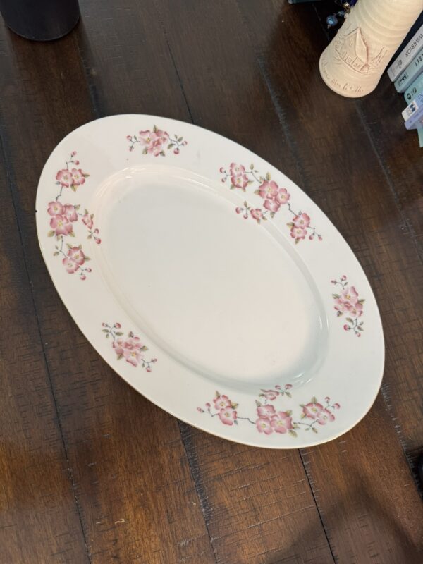 An oval white ceramic platter with a pattern of pink flowers and green leaves around the rim sits on a dark wooden table. Part of a cream-colored vase and some books are visible in the upper right corner of the image.