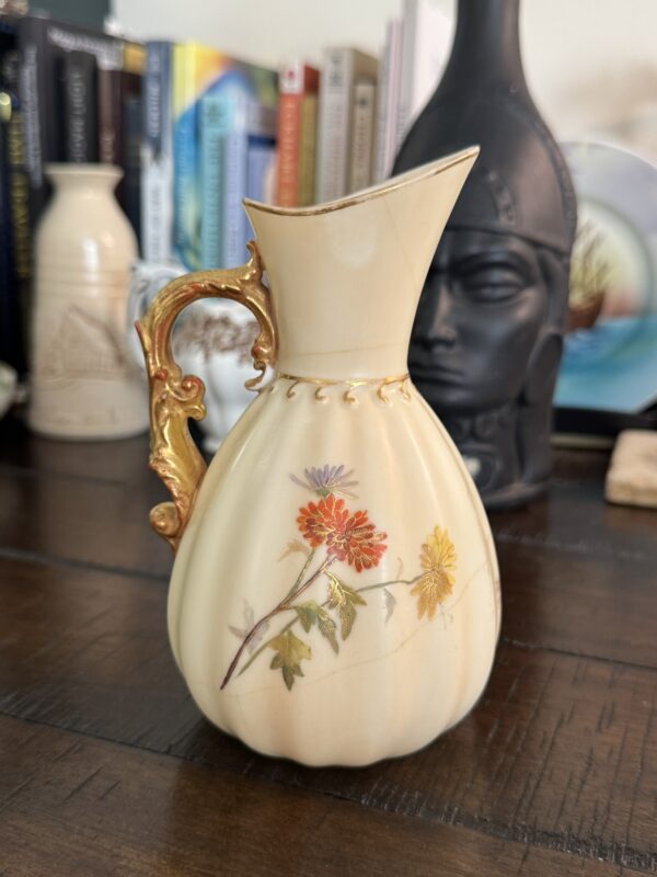 An ivory ceramic pitcher with a gold ornate handle and delicate red, purple, and yellow floral designs sits on a wooden table. Books and decorative items, including a helmet-shaped vase, are blurred in the background.