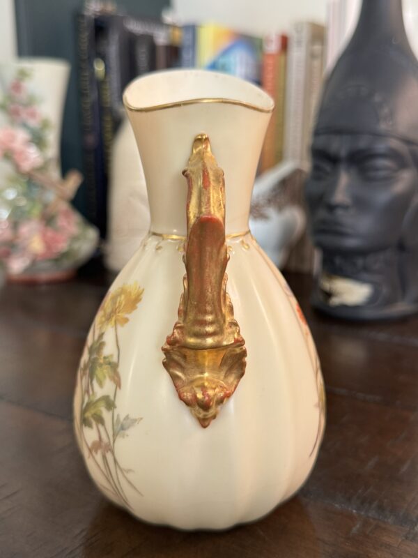 A cream-colored ceramic pitcher with yellow floral designs and a gold decorative handle sits on a wooden table. In the background are books, a floral teapot, and a dark sculpted bust with a helmet, slightly out of focus.