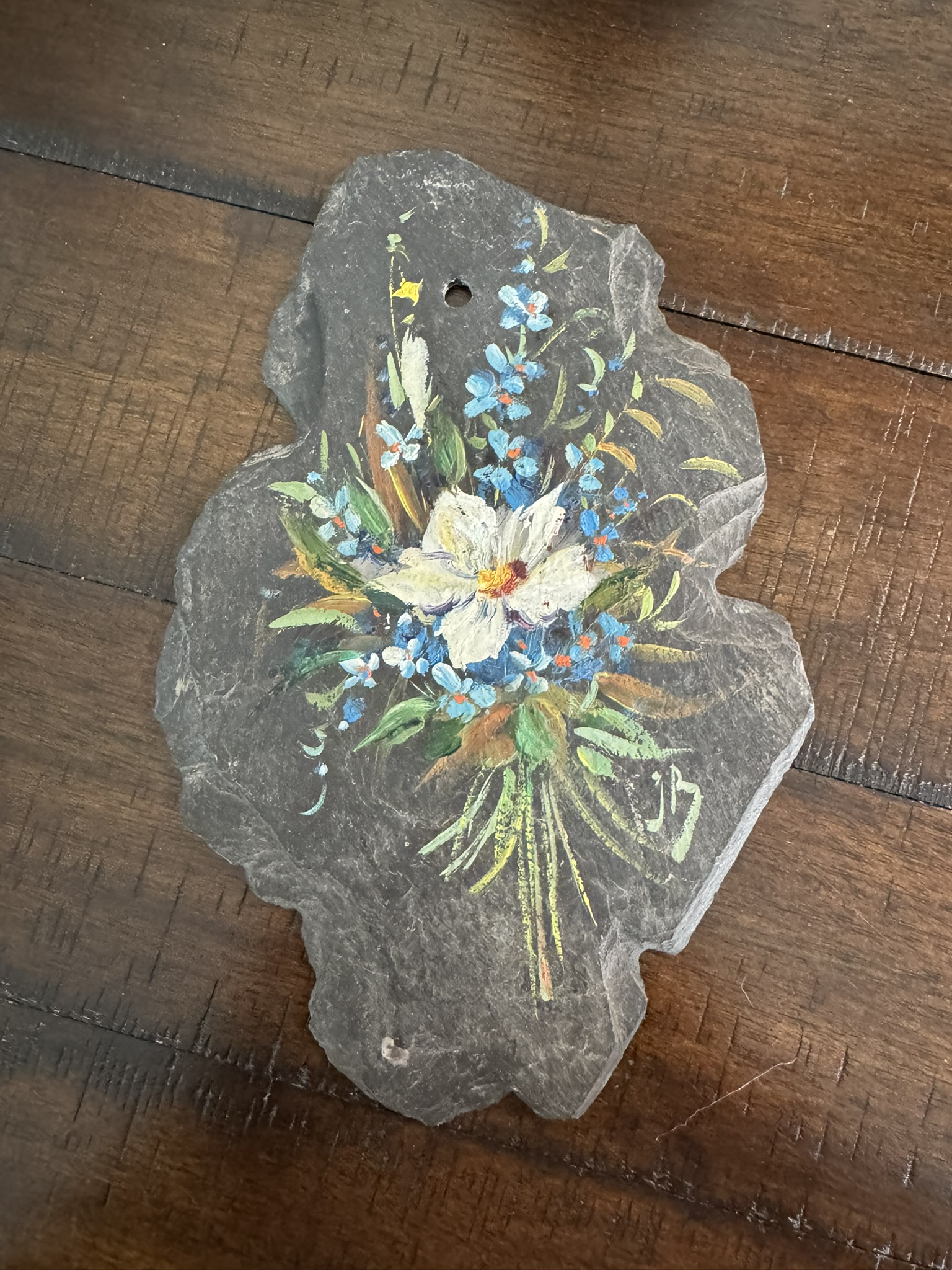 A piece of dark, irregularly shaped stone with a small hole at the top features a hand-painted bouquet of white and blue flowers with green stems and yellow accents, resting on a wooden surface.