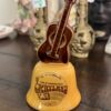 A ceramic souvenir bell from Nashville, Tennessee, featuring a brown guitar on top. The bell is yellow-orange with Grand Ole Opryland U.S.A. Home of American Music Nashville, Tennessee printed on the front. Decorative items are blurred in the background.