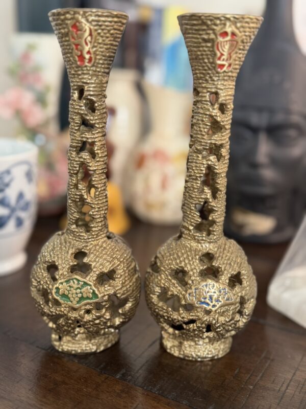 Two ornate, gold-colored vases with dragon designs and intricate cutouts stand side by side on a wooden table. Each vase features a colored accent—green on the left and blue on the right. Blurred decorative objects are visible in the background.