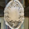 Oval ceramic platter with glossy white finish, featuring sculpted roses and floral accents around the rim. The platter rests on a colorful, distressed wood table, with visible chairs on either side. The floral details create an ornate, elegant border.