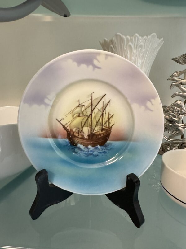 A decorative plate on a black stand features a painting of an old wooden sailing ship with tall masts and tan sails on blue ocean waves, under a sky with soft pink and purple clouds; white and silver decor is visible in the background.