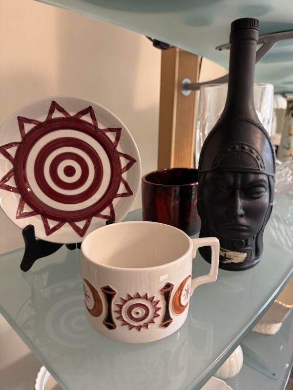 A white mug with red and brown geometric designs sits on a glass shelf. Behind it is a matching plate with a bold red sunburst and circle pattern. To the right is a matte black bottle shaped like a head, and a red glass cup is behind the mug.