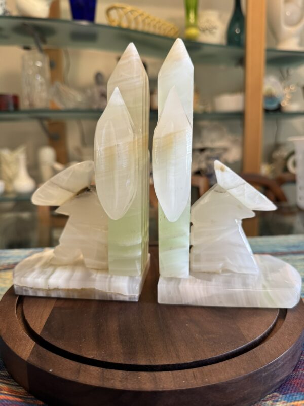 Two carved translucent onyx rabbit figures sit back-to-back, each facing outward, with tall crystal-like pillars rising behind them. They rest on square bases atop a round wooden plate. Shelves with glassware and ceramics appear in the blurred background.