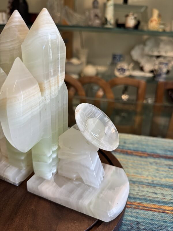 A carved, pale green and white onyx sculpture depicts pointed rocket shapes and a satellite dish on a wooden surface. In the background, glass shelves hold various small decorative objects, and wooden chairs are partially visible.