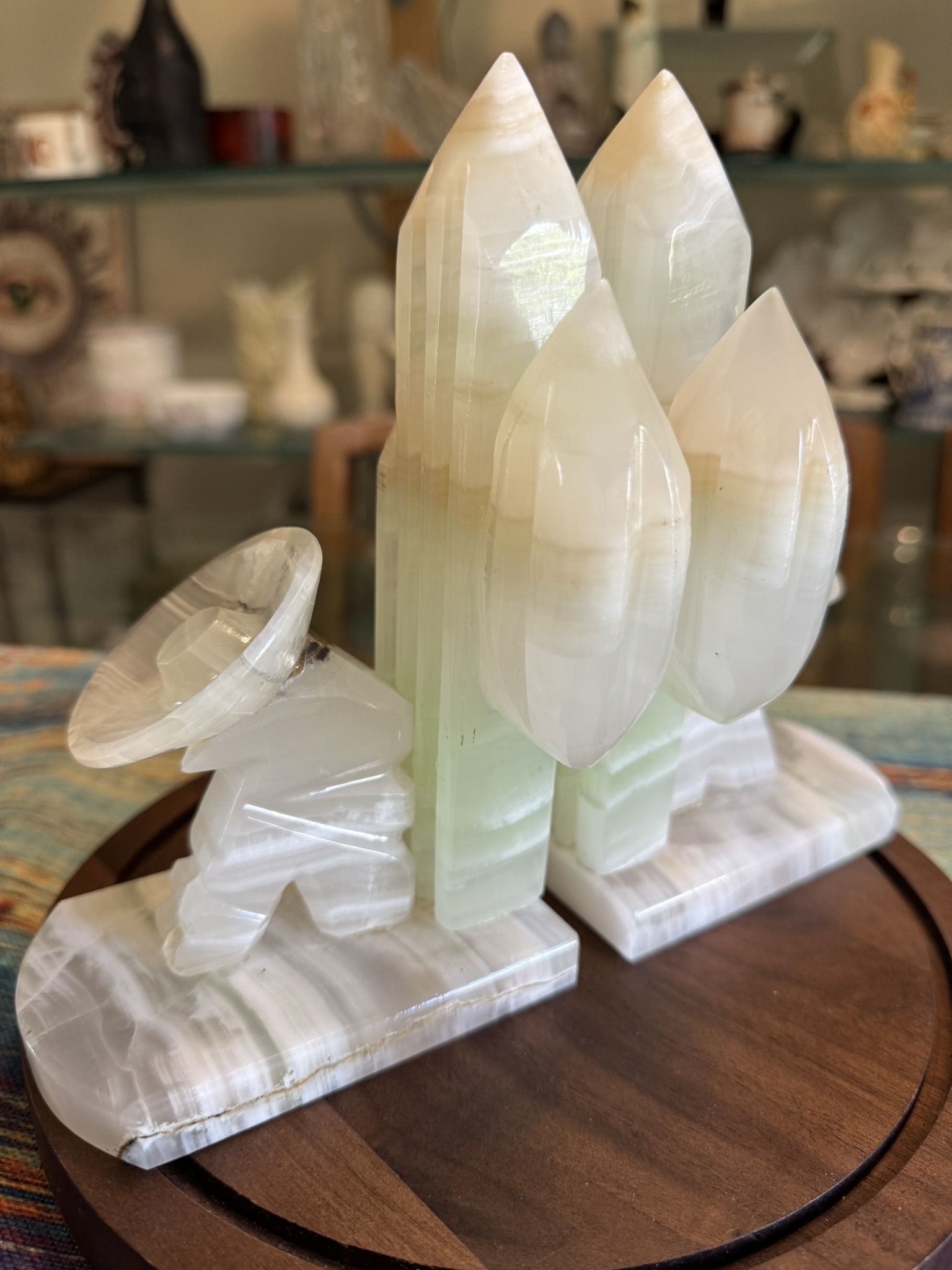 A polished onyx sculpture shows a rocket with three pointed boosters on a stand, next to a satellite dish aimed upward. Both rest on a round wooden base, with shelves of decorative objects blurred in the background.