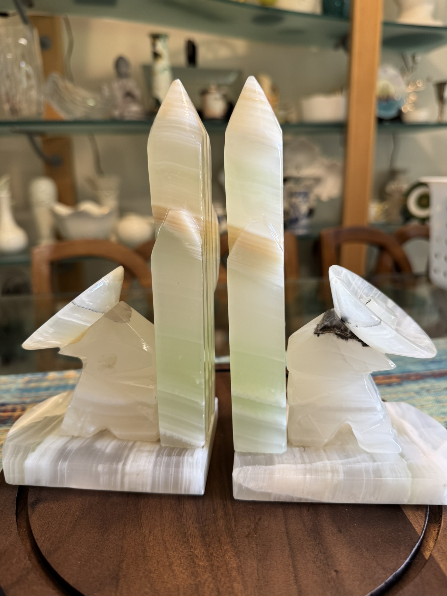 Two polished stone bookends shaped like wolves howling at tall, pointed obelisks. The wolves and obelisks are made of white and pale green onyx with brown veining. The background shows shelves with glassware and ceramic items in a home setting.