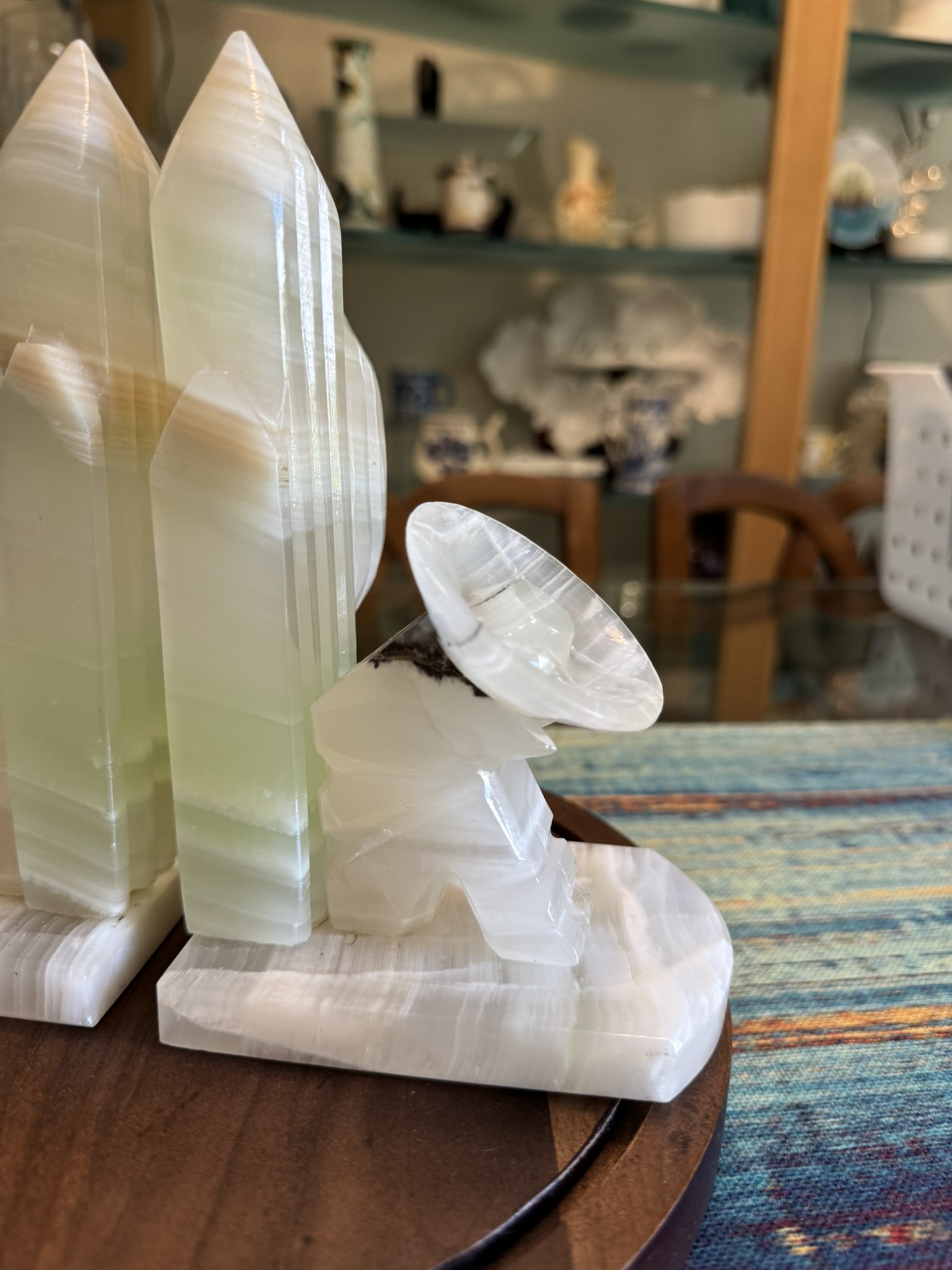 A carved onyx sculpture features two tall, pointed towers beside a small satellite dish, all on a rectangular base. The scene is set on a wooden table with shelves of ceramics and decorative items in the background.