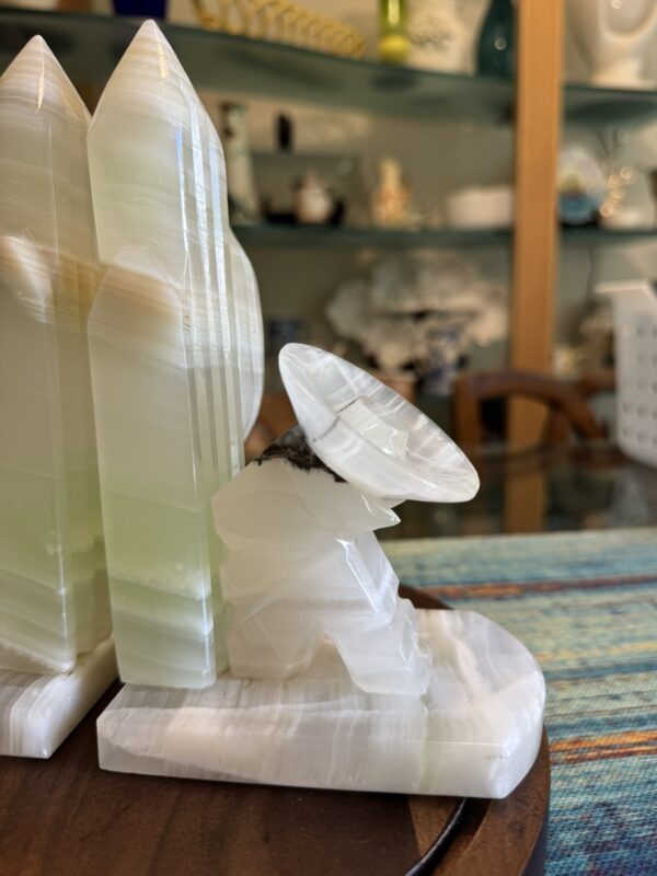 A close-up of a carved white stone figurine shaped like a bear holding a dish, likely made of onyx or marble, sits on a wooden surface. Behind it are tall, pointed stone shapes, with shelves of decorative items blurred in the background.