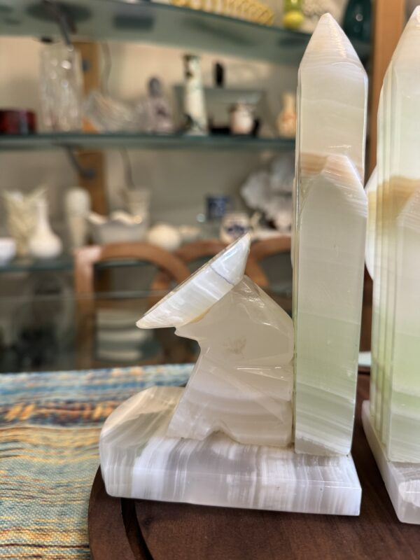 A carved onyx bookend shaped like a howling wolf next to a tall obelisk stands on a table. The stone is cream and light green with natural banding. In the background, glass shelves display various decorative items and glassware.