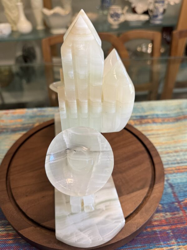 A polished white onyx sculpture with three pointed towers and a circular disc in front, mounted on a wooden base. The background shows a multicolored woven tablecloth and a glass display cabinet with various decorative objects.